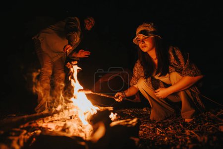 Grupo de amigos que experimentan la alegría de estar juntos mientras preparan comida sobre una fogata al aire libre por la noche junto a un lago.