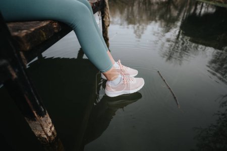 Un moment paisible en tant que femme en tenue de sport profite d'une pause au bord de l'eau, mettant en valeur ses baskets et l'environnement serein.
