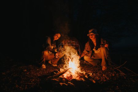 Two friends experiencing the warmth and joy of a campfire outdoors at night, preparing food and cherishing their togetherness by a serene lake.