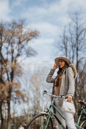 Modische junge Frau mit schickem Hut steht neben ihrem Fahrrad und umarmt die frische, kühle Luft eines sonnigen Herbsttages im Park.