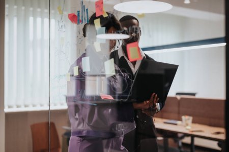 Ein fokussierter afrikanischer Mann und eine kaukasische Frau nehmen an einer strategischen Planungssitzung teil und verwenden einen Laptop und klebrige Notizen an einer Glaswand in einem hell erleuchteten Büro..