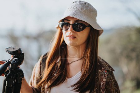 Une jeune femme confiante ajuste sa caméra sur un trépied à l'extérieur. Matériel photo professionnel, lumière naturelle et arrière-plan scénique améliorent le cadre.