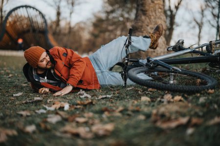 Lässiger junger Mann im Hipster-Stil stürzt in einem Stadtpark von seinem Fahrrad auf den Rasen.