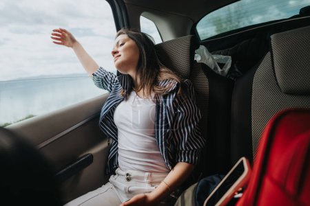 Captured inside a car, this image shows a young woman happily stretching her arm out, feeling the breeze as she enjoys a scenic road trip.