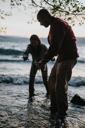 Dans une scène de coucher de soleil évocatrice, un homme assiste une femme dans le contexte des vagues orageuses du lac, symbolisant le soutien et les soins pendant les périodes difficiles.