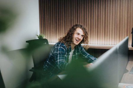 Captured in an office setting, a young male employee with curly hair smiles engagingly at a colleague, conveying a friendly, collaborative spirit among workers.