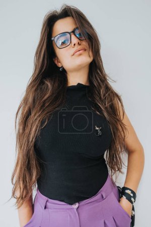 A stylish young woman stands confidently in a black top and purple pants, her brown hair flowing and glasses on.