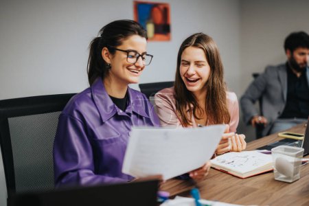Two young female entrepreneurs laugh while reviewing project charts at a lively startup meeting, showcasing teamwork and productivity in a multicultural office environment.