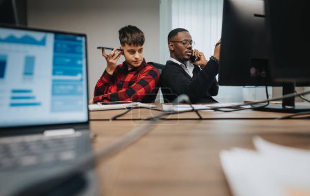 Zwei männliche Kollegen, einer telefoniert, arbeiten in einem Büro zusammen, umgeben von Computern und Geschäftsunterlagen..
