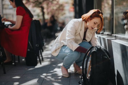 Konzentrierte junge Unternehmerin auf der Suche nach ihrem Rucksack im Freien mit Café-Kulisse im Hintergrund.