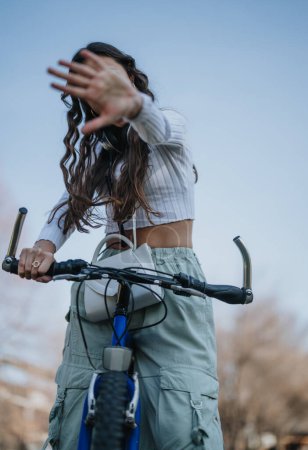 Lässige junge Frau auf dem Fahrrad gestikuliert, um nicht an einem sonnigen Tag in einer städtischen Umgebung fotografiert zu werden.