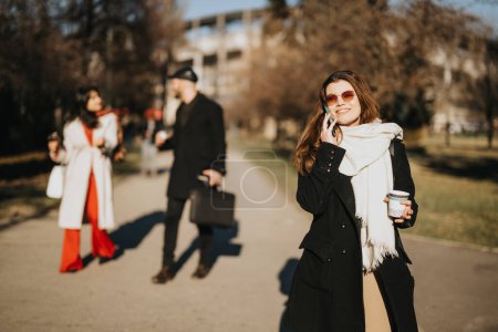 In einem schönen Outdoor-Ambiente führt eine fröhliche junge Geschäftsfrau an einem hellen Wintertag ein Telefongespräch, im Hintergrund diskutieren Kollegen..