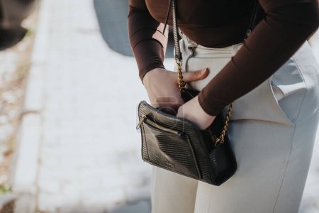 Elegant businesswoman holding a luxurious black designer handbag, dressed in a sophisticated outfit, showcasing elements of professional fashion and style in an urban setting.