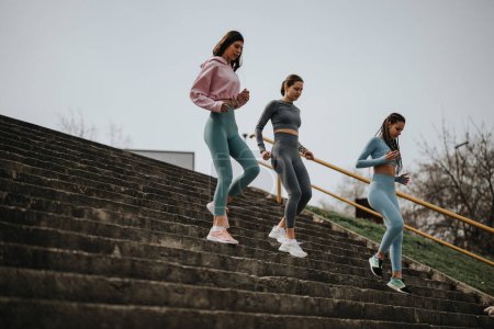 Three athletic women in workout gear descend a long staircase while exercising in an urban outdoor setting, promoting health and fitness.