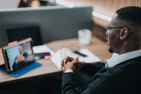 Hombre de negocios afroamericano participando en una reunión de negocios virtual, concentrándose en una discusión con un colega visible en una pantalla de tableta.