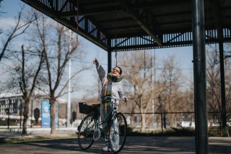 Ältere, glückliche Dame mit Fahrrad genießt einen sonnigen Tag im Freien und zeigt auf etwas Interessantes oben in einem Stadtpark.