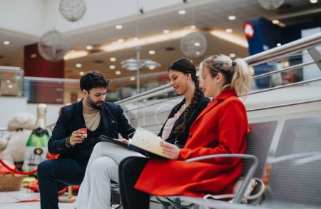 Three business colleagues engage in a discussion with paperwork and coffee at a shopping mall lounge area.