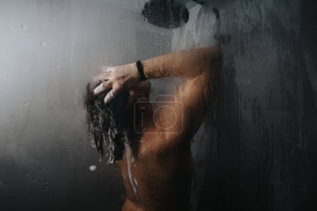 Moody and artistic image of a man taking a shower, hand in hair, seen behind a wet glass pane.