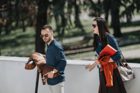 Un homme et une femme en tenue de travail décontractée se tiennent à l'extérieur, semblant concentrés et en déplacement avec des objets personnels à la main.