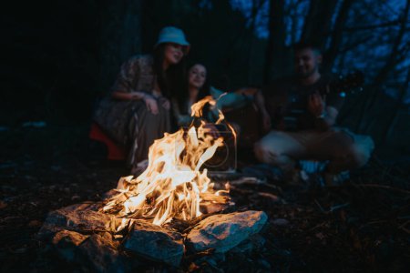 Three friends gather around a blazing campfire in the forest, sharing stories and playing guitar under the evening sky.