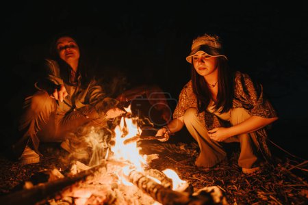 Two friends toasting marshmallows over a campfire at night beside a lake, sharing happy moments in nature.