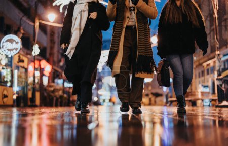 Three people walking on a city street at night, reflecting lights on wet pavement, urban life