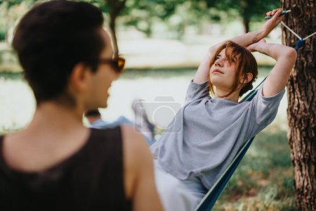 Eine Frau liegt in einer Hängematte mit einem jungen Mann in der Nähe, beide genießen einen ruhigen Moment in einem üppigen Park an einem klaren Tag.