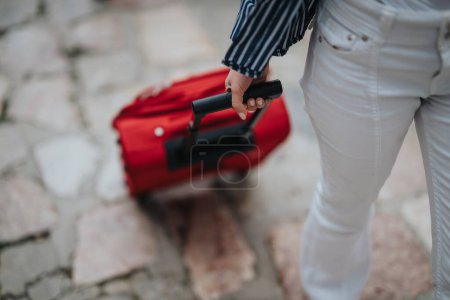 In a sunny town, a young tourist enjoys a leisurely stroll, pulling a red suitcase along the charming cobblestone street.
