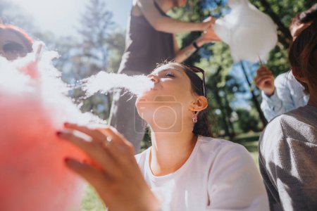 Summer joy as a young woman savors cotton candy while relaxing in a park with sunlit trees and friends around.