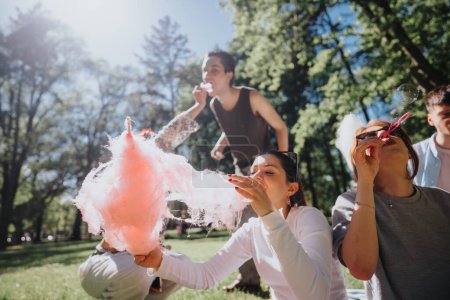 A joyful group of young adults enjoying cotton candy and blowing bubbles on a sunny day in a green park, embodying freedom and leisure.