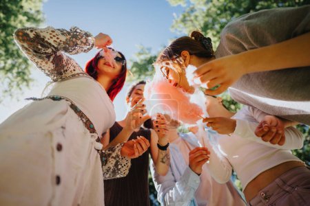 Grupo de amigos alegres comiendo algodón de azúcar bajo la sombra de los árboles en un parque soleado, que representa la felicidad y el ocio.