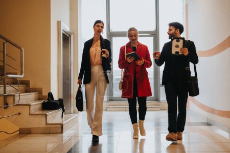 Three stylish friends casually walking with coffee cups and modern devices in a bright corridor.