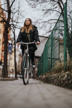 A determined young woman on a bicycle pedals along a city sidewalk, showcasing an active urban lifestyle during the fall season.