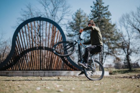 Mitten im Geschehen führt ein Mann einen Wheelie auf einem Fahrrad vor und zeigt Geschicklichkeit und Gleichgewicht gegen einen Park mit moderner Brückenstruktur.