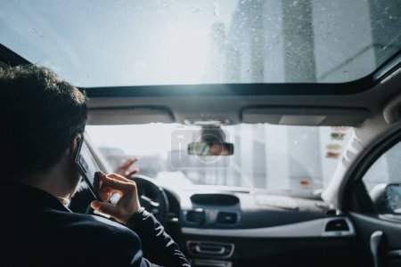 Vue arrière d'un homme en costume faisant un appel téléphonique sur le siège arrière d'une voiture, avec une lumière du jour brillante venant par la fenêtre.