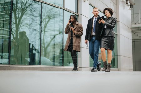 Drei multiethnische Geschäftspartner in trendiger Oberbekleidung gehen vor einem Bürogebäude spazieren und führen ein ernsthaftes Geschäftsgespräch.