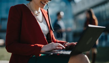 Selbstbewusste Geschäftsfrau im roten Blazer, die an einem Laptop mit Kopfhörer in sonnigem Outdoor-Ambiente arbeitet und mobiles Büro und Flexibilität verkörpert.