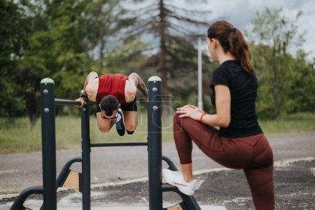 A focused man mastering calisthenics by doing an invert on horizontal bars while a woman in active wear prepares nearby, set against a lush park backdrop.