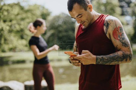 A man with tattoos checks his phone as his female jogging partner stretches in the background in a lush, green park.