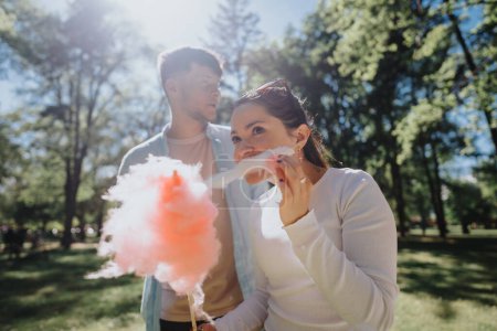 Ein fröhliches junges Paar genießt es, an einem strahlend sonnigen Tag gemeinsam in einem üppigen Park rosa Zuckerwatte zu essen und einen Moment der Freude zu teilen.