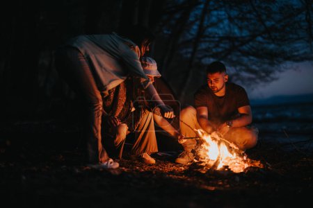 Groupe d'amis préparant la nourriture sur un feu de camp au bord du lac, embrassant ensemble dans la nature au crépuscule.