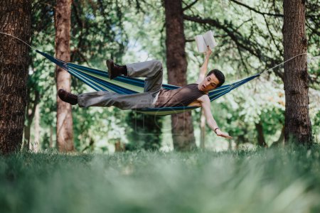 Ein fröhlicher junger Mann balanciert in einer Hängematte, während er ein Buch liest, und zeigt Ausgeglichenheit und Entspannung inmitten einer sonnigen Parklandschaft.