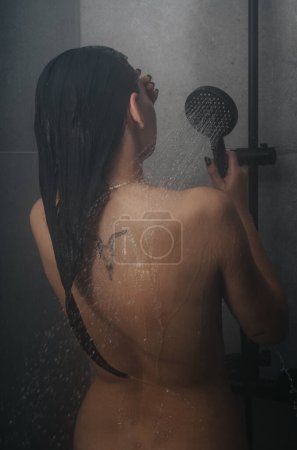 Back view of a young female using a shower head, water cascading down her skin in a serene bathroom atmosphere.