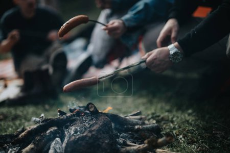 Close-up of a sausage being roasted over a campfire, with friends gathered in a forest setting.