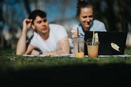 Affectionate student couple enjoying a study session in the park with cold beverages and a notebook.