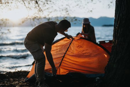 Two friends enjoy their weekend outdoors as they set up a camping tent by a beautiful lake at sunset, embracing relaxation and togetherness.