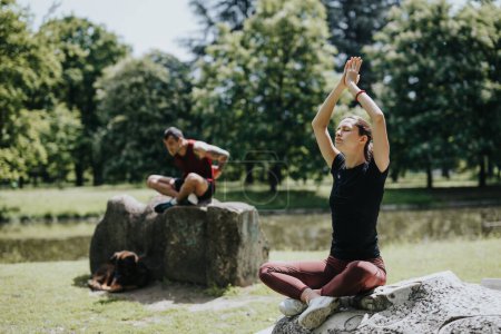 Un hombre y una mujer, posiblemente amigos, participan en rutinas de fitness en un parque escénico la mujer practica yoga mientras el hombre descansa cerca.