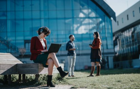 Eine fokussierte Geschäftsfrau mit Kopfhörer benutzt einen Laptop, während sie im Freien auf einer Bank sitzt und sich im Hintergrund mit zwei Kollegen unterhält.