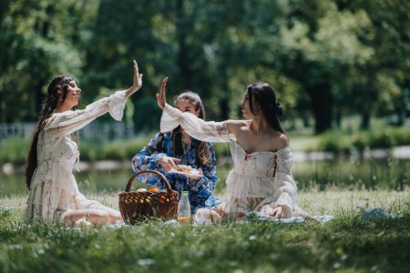 Image captures three young women in floral dresses having fun during a picnic in a lush green park, with a basket of food and happy expressions.