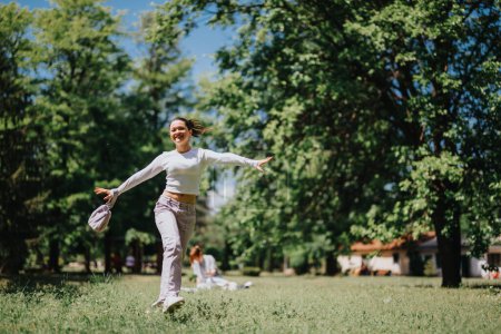 A happy young woman spreads her arms wide as she walks carefree through a sunny, green park, embodying joy and freedom.